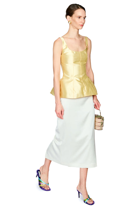 A model is shown from the head to toe wearing a sleeveless yellow peplum top and a light cream midi skirt, holding a small woven clutch and wearing colorful high-heeled sandals against a plain white background.