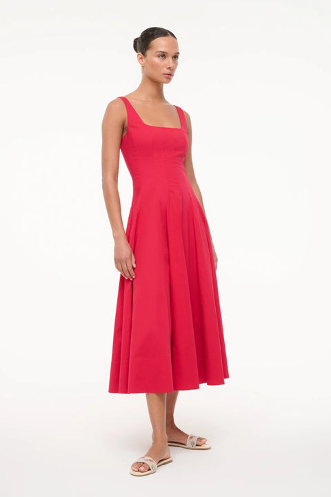 Side angle of a woman wearing a red sleeveless midi dress that features a square neck, corset-style paneling, and an A-line skirt, paired with white sandals. 