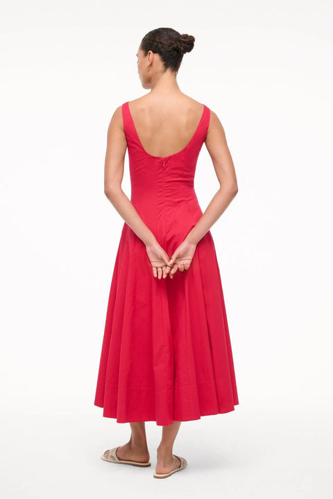 Back angle of a woman wearing a red sleeveless midi dress that features a scoop back, corset-style paneling, and an A-line skirt, paired with white sandals. 