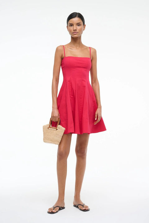 Woman wearing a red mini dress that features spaghetti straps and a straight neckline, corset style paneling, and an A-line skirt, paired with a crochet handbag and black sandals. 