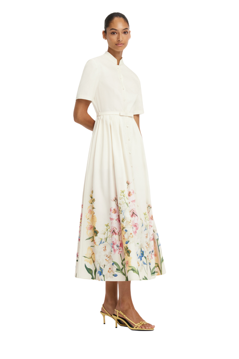 A model wearing a white midi dress with a floral print concentrated on the lower skirt, styled with gold heeled sandals against a plain white background.