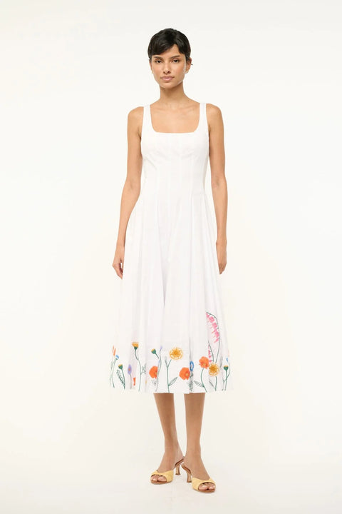Woman wearing a sleeveless white midi dress that features a square neck, corset-style paneling, and colorful floral embroidery at the bottom. 