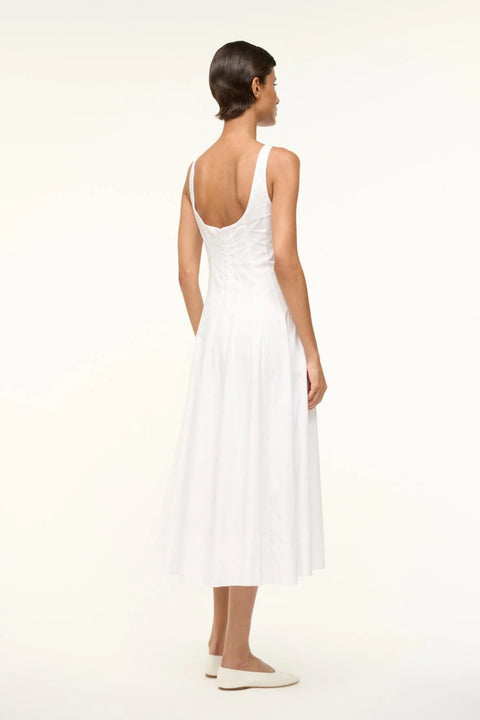 Back view of a woman wearing a sleeveless white midi dress that features a square neck, corset-style paneling, and an A-line skirt, paired with white flats. 