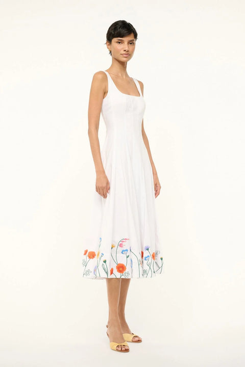 Side view of a woman wearing a sleeveless white midi dress that features a square neck, corset-style paneling, and colorful floral embroidery at the bottom. 