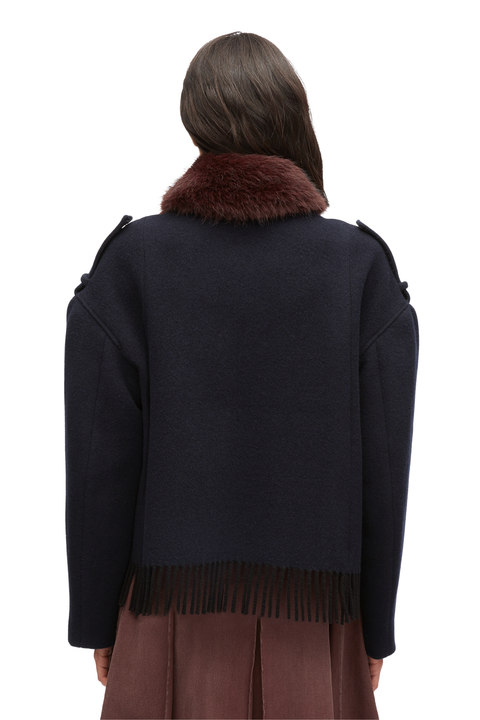 A model shown from the back wearing a dark navy blue cropped sweater with a brown faux-fur collar and fringe trim at the hem, paired with a brown skirt against a plain white background.