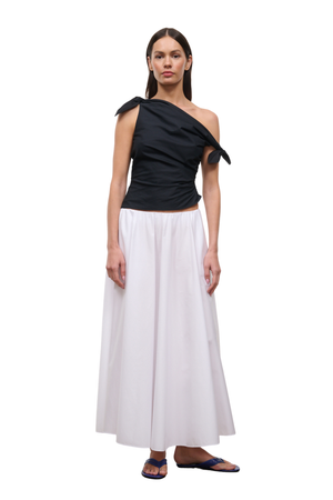 A woman modeling a two-piece outfit consisting of a black one-shoulder draped top with tie details at the shoulders and a long white skirt, paired with dark blue flip-flop sandals, against a plain white background.