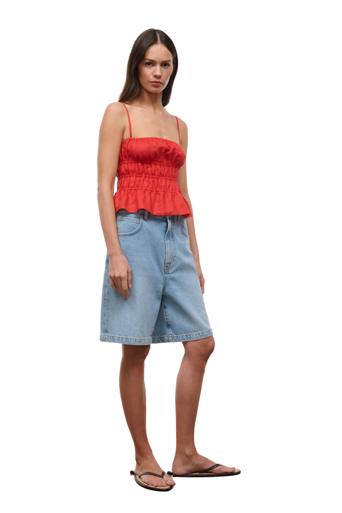 A woman modeling a bright red ruffled spaghetti-strap camisole top paired with light-wash denim shorts and dark flip-flop sandals, set against a plain white background.