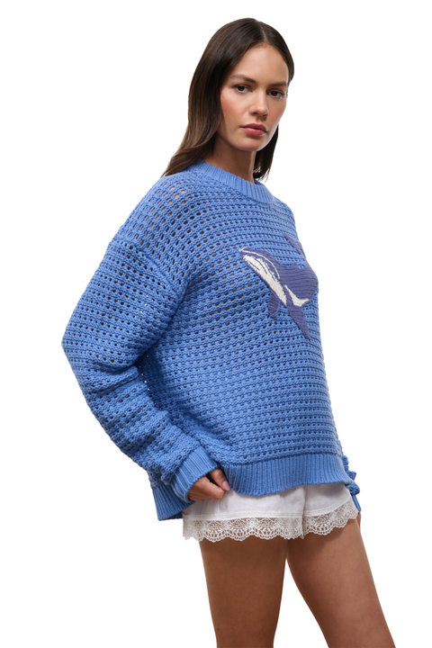 A woman wearing a chunky blue crocheted sweater with a white and blue whale graphic on the chest, paired with white lace hem shorts, against a plain white background.