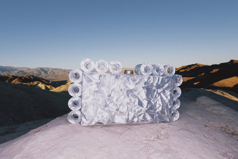 A rectangular acrylic clutch that features a silver foil design and matching open circles of silver-foiled acrylic lining the shape of the clutch, set on a ledge with mountains in the background. 