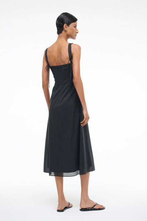 Back-facing woman wearing a black Victorian-inspired midi dress that features lace straps, corset-style detailing, and buttons down the front, paired with black sandals. 