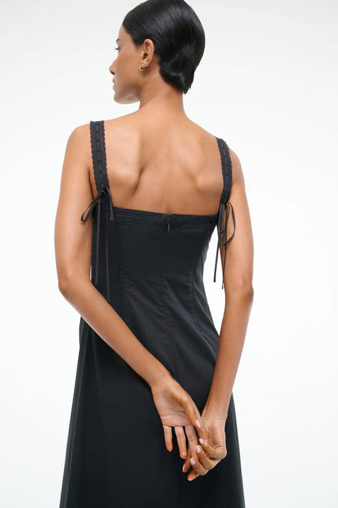 Close up of a back-facing woman wearing a black Victorian-inspired midi dress that features lace straps, corset-style detailing, and ribbon bows on the back. 