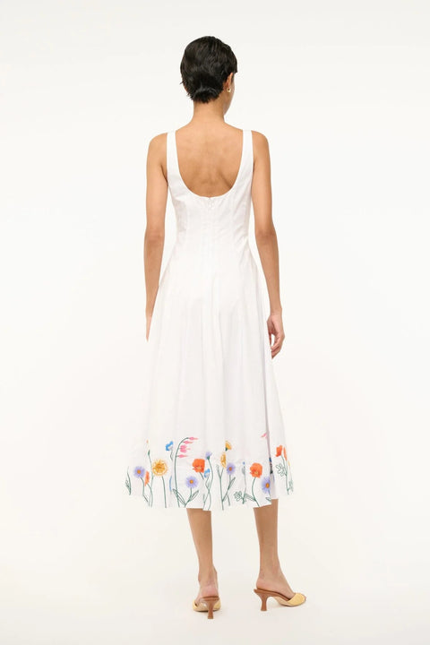 Back view of a woman wearing a sleeveless white midi dress that features a square neck, corset-style paneling, and colorful floral embroidery at the bottom.
