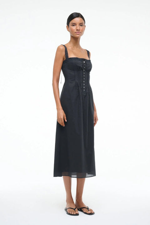 Side-facing woman wearing a black Victorian-inspired midi dress that features lace straps, corset-style detailing, and buttons down the front, paired with black sandals. 