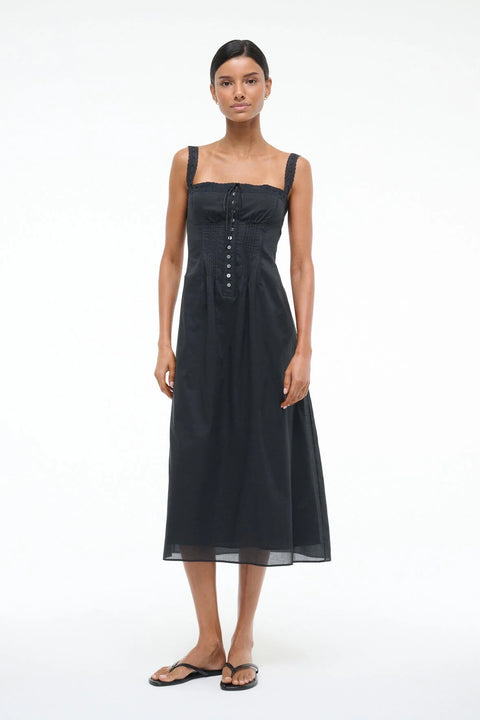 Woman wearing a black Victorian-inspired midi dress that features lace straps, corset-style detailing, and buttons down the front, paired with black sandals. 