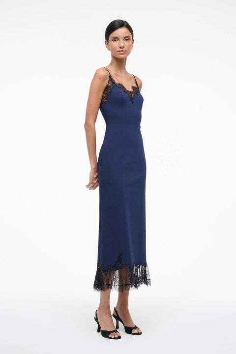Side-facing woman wearing a navy midi slip dress that features black lace trim along the neckline and hem, paired with black kitten heels. 