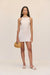 Front-facing woman wearing a cream linen mini dress that features a unique, threaded halter neckline, paired with cream sandals and a woven handbag. 