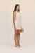 Side-facing woman woman wearing a cream linen mini dress that features a unique, threaded halter neckline, paired with cream sandals and a woven handbag. 