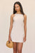 Close up of a front-facing woman wearing a cream linen mini dress that features a unique alter neckline, paired with a woven handbag. 