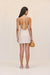 Back-facing woman wearing a cream linen mini dress that features thick straps which tie into a halter neck, as well as an open back, paired with cream sandals and a woven handbag. 