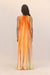 Back view of a woman wearing an orange ombre maxi dress that features a high neck and ribbons of ilk woven to form cutouts on the shoulders before cascading toward the bottom of the dress. 