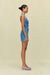 Side view of a woman wearing a blue denim mini dress with a halter neck and torso cutouts, paired with chunky gold earrings and denim mules. 