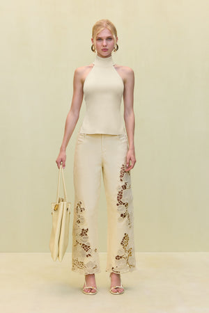 Woman wearing a pair of light yellow, mid rise pants that feature a wide leg silhouette with open floral embroidery details on the front and side of the legs, paired with a light yellow top and tote. 