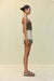 Side view of a woman wearing an olive green knit tank top that features a gold-toned chain detail for straps and across the neckline, paired with cream shorts and sandals. 