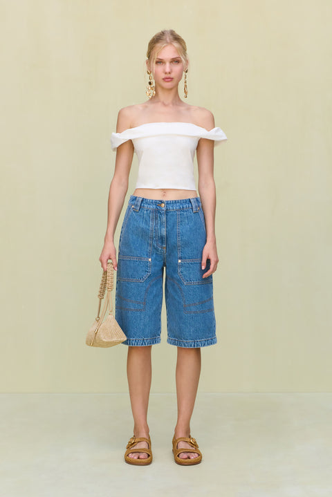 Woman wearing a white, off-the-shoulder crop top, paired with long blue jean shorts, sandals, and gold statement earrings. 