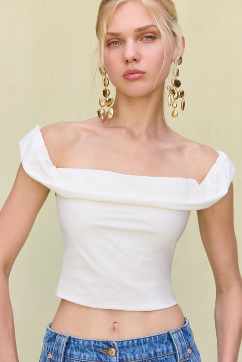 Close up of a woman wearing a white, off-the-shoulder crop top, paired with long blue jean shorts and gold statement earrings. 