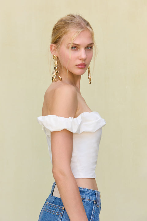 Side view of a woman wearing a white, off-the-shoulder crop top , paired with long blue jean shorts and gold statement earrings. 