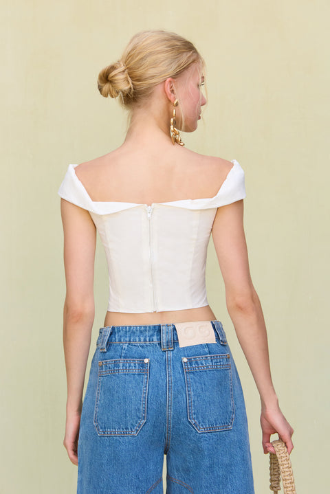 Back view of a woman wearing a white, off-the-shoulder crop top , paired with long blue jean shorts, sandals, and gold statement earrings. 