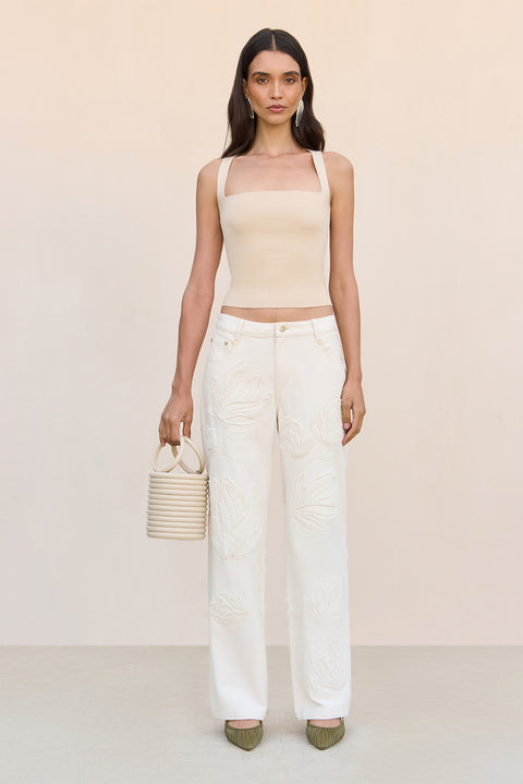 Woman wearing a beige top and white pants holding a straw bag on a white background standing centered.