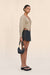 Side-facing woman wearing a structured beige cardigan and black skirt holding a black handbag on a beige background