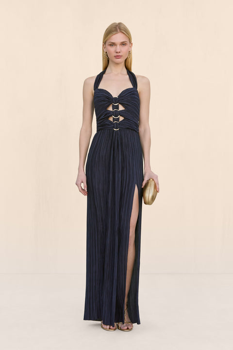 Woman wearing a navy plisse halter gown that features a sweetheart neckline, cutouts at the chest fasten by gold rings, and a high leg slit, paired with gold accessories. 