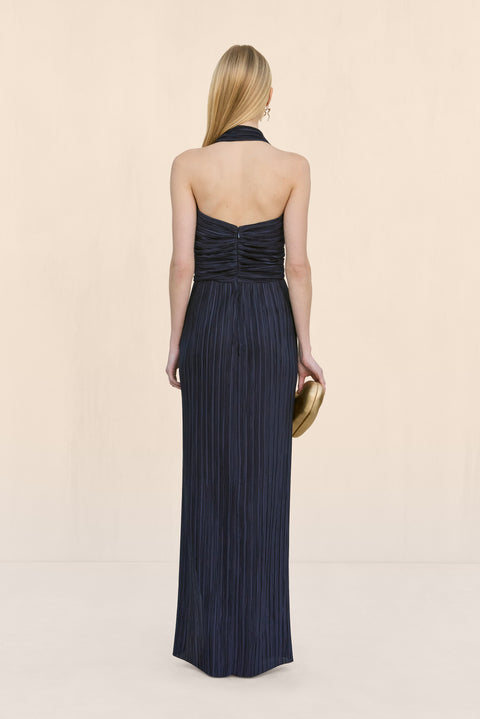 Back view of a a woman wearing a navy plisse gown with a halter neck and open back, paired with a gold clutch. 