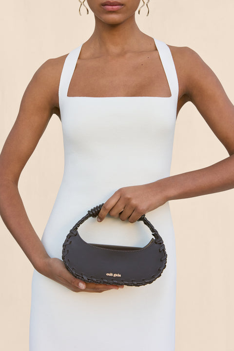 Woman wearing a sleeveless, white mini dress that features a square neckline and holding a black handbag. 