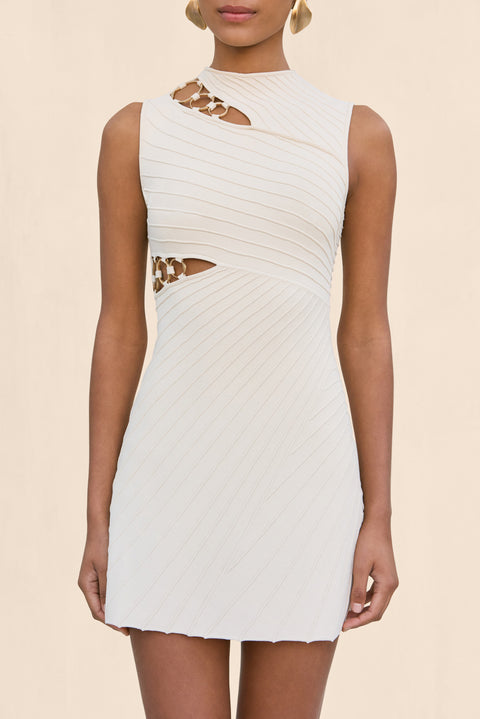 Close up of a woman wearing a cream, sleeveless mini dress that features cutout details with rings at the shoulder and ribcage. 