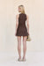 Back view of a woman wearing a sleeveless, brown knit dress that features a center zipper down the back, paired with a tan handbag and tan sandals. 