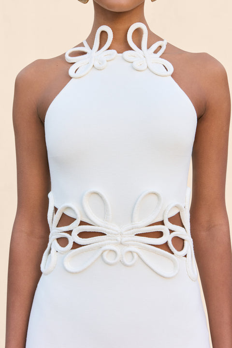 Close up of a woman wearing a white knit midi dress that features cutouts at the waist with a floral overlay and a floral applique at the neckline. 