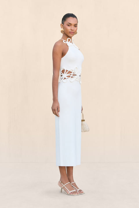 Side-facing woman wearing a white knit midi dress that features cutouts at the waist with a floral overlay and a floral applique at the neckline. 