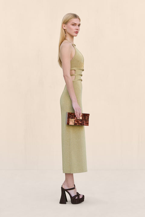 Side view of a woman wearing a gold knit midi dress that features a halter neck, deep V, four buckle details across the bodice, and an open back, paired with black accessories. 