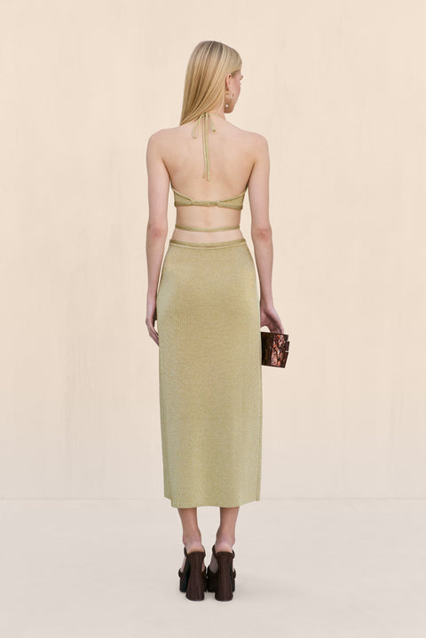 Back-facing woman wearing a gold knit midi dress that features a halter neck and open back secured by two straps across the back, paired with black heels. 