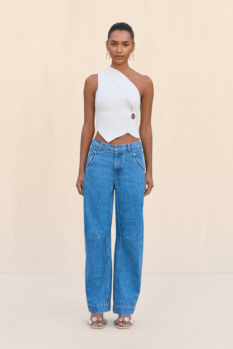 Woman wearing a white knit off the shoulder crop top that features a cutout at the rib cage and an asymmetrical hem, paired with blue wide leg jeans.