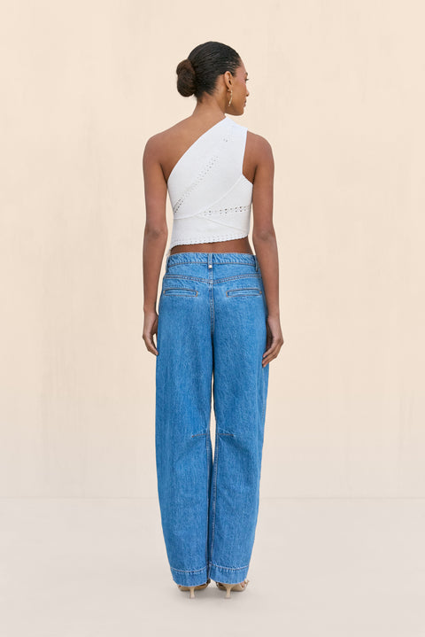 Back-facing woman wearing a white knit off the shoulder crop top that features a cutout at the rib cage and an asymmetrical hem, paired with blue wide leg jeans.