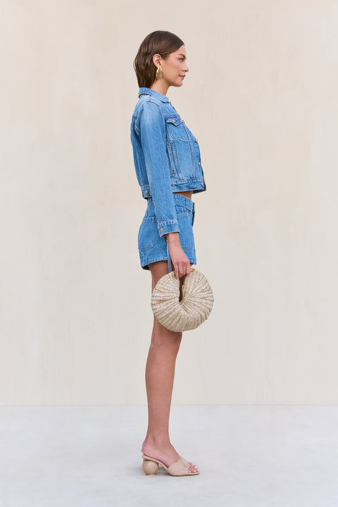 Side view of a woman wearing blue denim cargo shorts paired with a matching denim jacked and a woven handbag. 