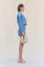 Side view of a woman wearing blue denim cargo shorts paired with a matching denim jacked and a woven handbag. 