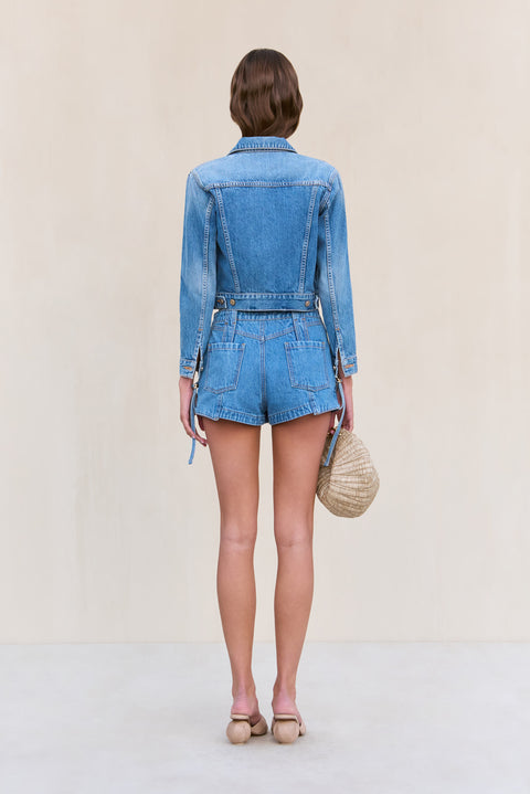 Back view of a woman wearing blue denim cargo shorts paired with a matching denim jacket and a woven handbag. 