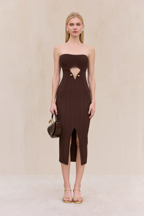 Woman wearing a brown knit, strapless midi dress with cut-out design at the bust featuring gold hardware, and a front slit, paired with gold shoes. 