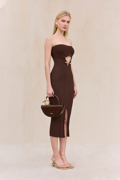 Side view of a woman wearing a brown knit, strapless midi dress with cut-out design at the bust featuring gold hardware, and a front slit, paired with gold shoes. 