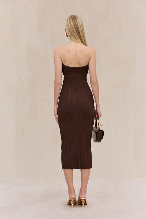 Back-facing woman wearing a brown knit, strapless midi dress, paired with gold heels and a brown handbag. 
